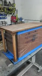 Wooden cabinet on a workbench, wrapped in plastic and blue tape, in a cluttered workshop.