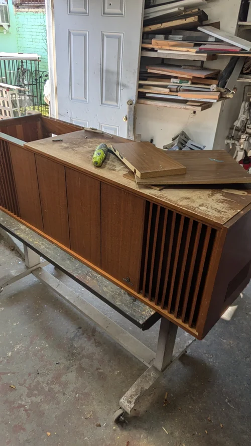 Wooden cabinet on a metal stand, being refurbished with tools and wood pieces nearby.